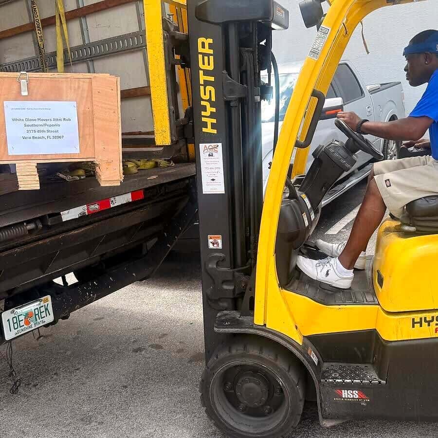A forklift sets a labeled wooden crate on the liftgate of a box truck with a Florida plate.
