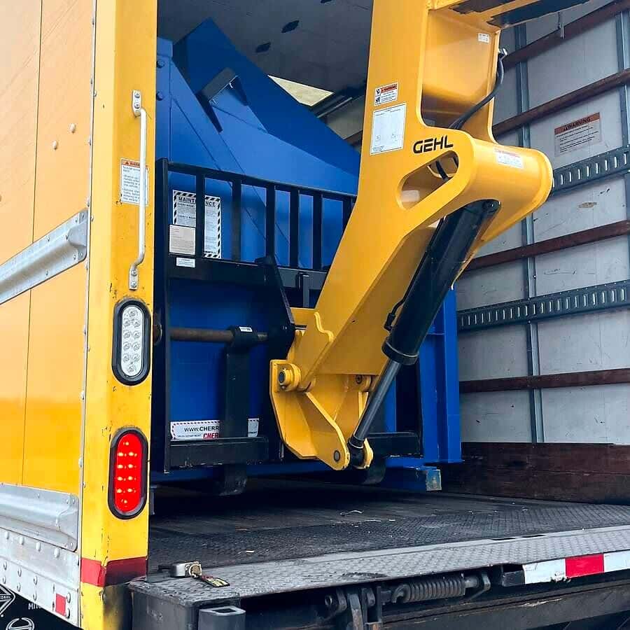A telehandler places a heavy piece of blue equipment into the box truck over the liftgate.