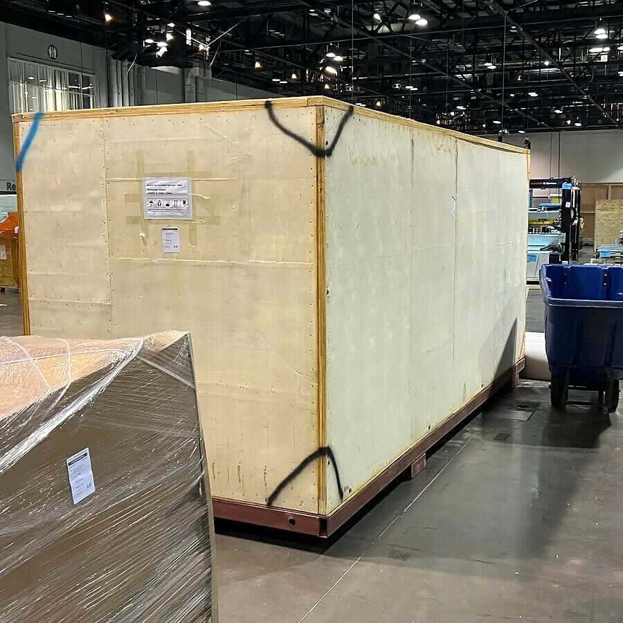 A very large wooden crate and a wrapped pallet inside a large exhibition or warehouse hall.